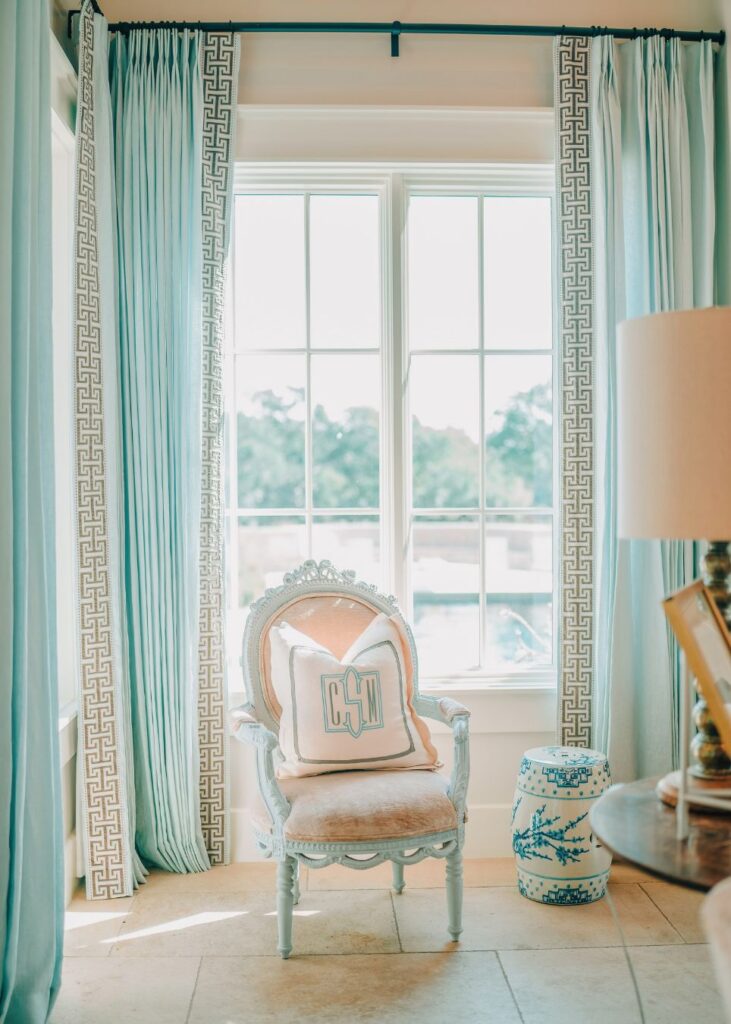 A sunlit room styled by an interior decorator in Boerne features tall aqua drapes, a garden-view window, an ornate light blue chair with monogrammed pillows, a white and blue ceramic stool, and a lamp partly visible on the right.