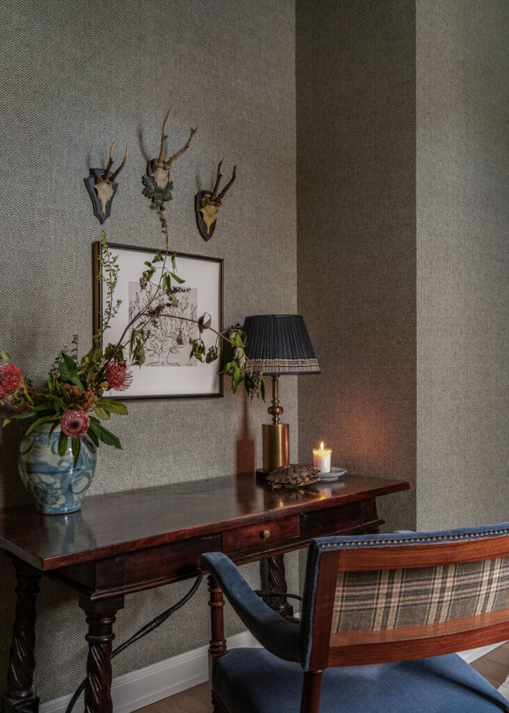 A cozy study corner with a wooden desk, blue upholstered chair, lamp, lit candle, vase of flowers, framed artwork, and three mounted antelope skulls on a textured gray wall.