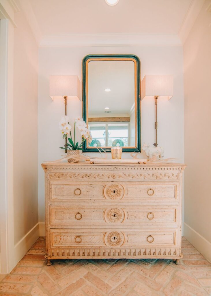 A light wood dresser with ornate carvings stands below a large framed mirror, flanked by wall-mounted lamps. Styled by an interior decorator Boerne, a potted white orchid and decor add elegance to this bright, well-lit hallway.