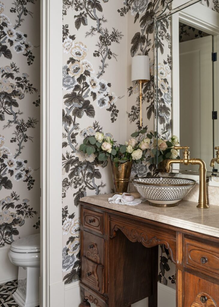 A bathroom curated by an interior designer in Boerne, TX, features floral wallpaper, a wooden vanity with marble countertop, patterned vessel sink with gold faucet, wall mirror, white roses in a vase, and a glimpse of the toilet in the corner.