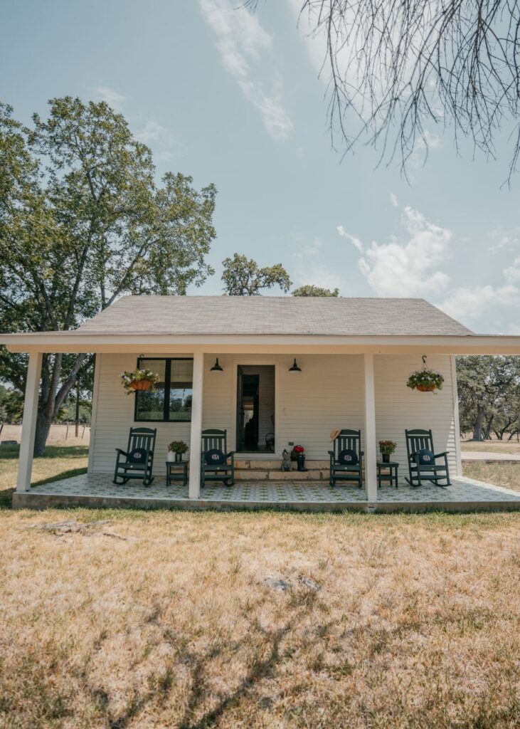 A small white house with a front porch featuring four dark rocking chairs, potted plants, and hanging flower baskets—beautifully styled like an interior design company in Boerne, TX—surrounded by trees and dry grass under a partly cloudy sky.