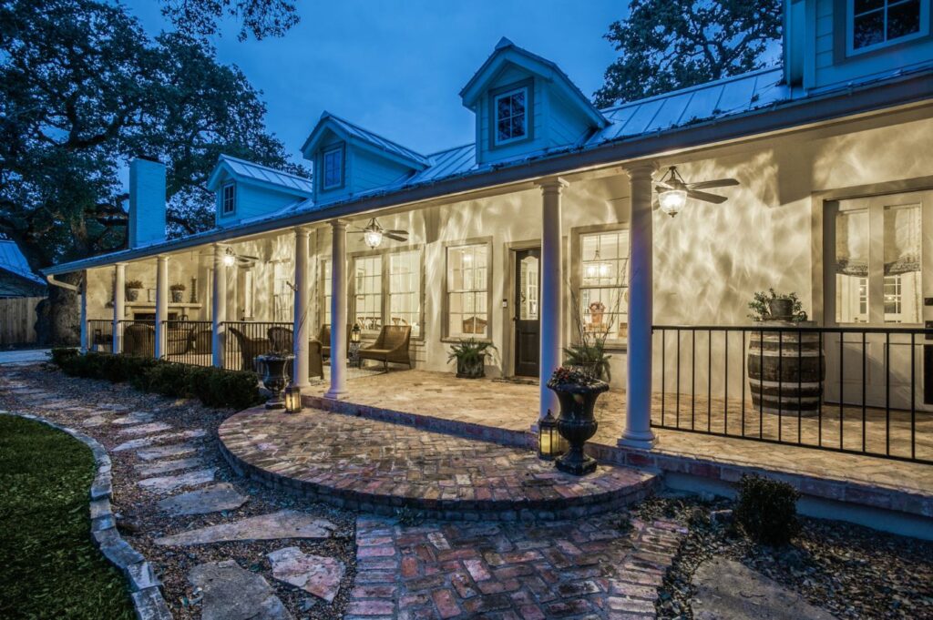 A charming, well-lit house with a wide front porch and elegant columns, captured at dusk. As an interior design company in Boerne, TX, we appreciate how thoughtful landscaping and outdoor furnishings enhance every welcoming entrance.