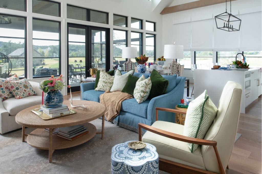 Bright, modern living room with large windows, a blue sofa with green pillows, a round wooden coffee table, and patterned armchairs. Styled by an interior designer Boerne TX, natural light fills this cozy, inviting space with an open kitchen in the background.