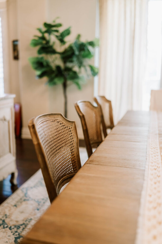 Three wooden chairs with woven cane backs are lined up next to a long dining table. A leafy plant is in the background, and light streams through sheer curtains—an inviting scene perfected by an interior design company Boerne TX residents trust.