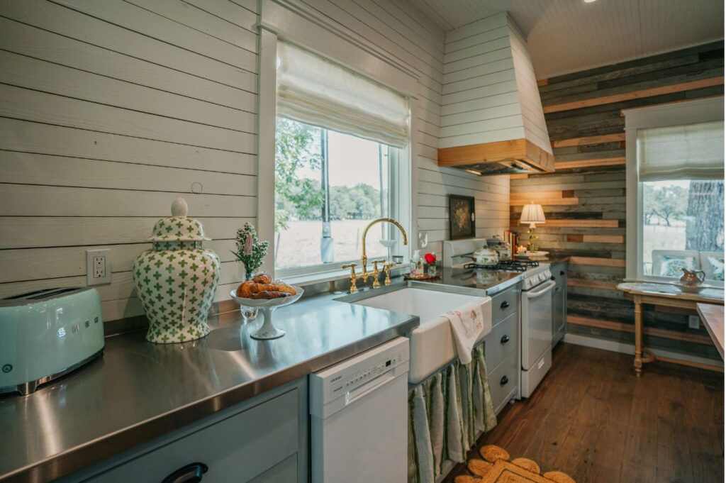 A cozy kitchen by an interior designer Boerne TX, featuring stainless steel countertops, a farmhouse sink, mint green toaster, decorative jars, and wooden accents. Large windows fill the space with natural light and views of trees outside.