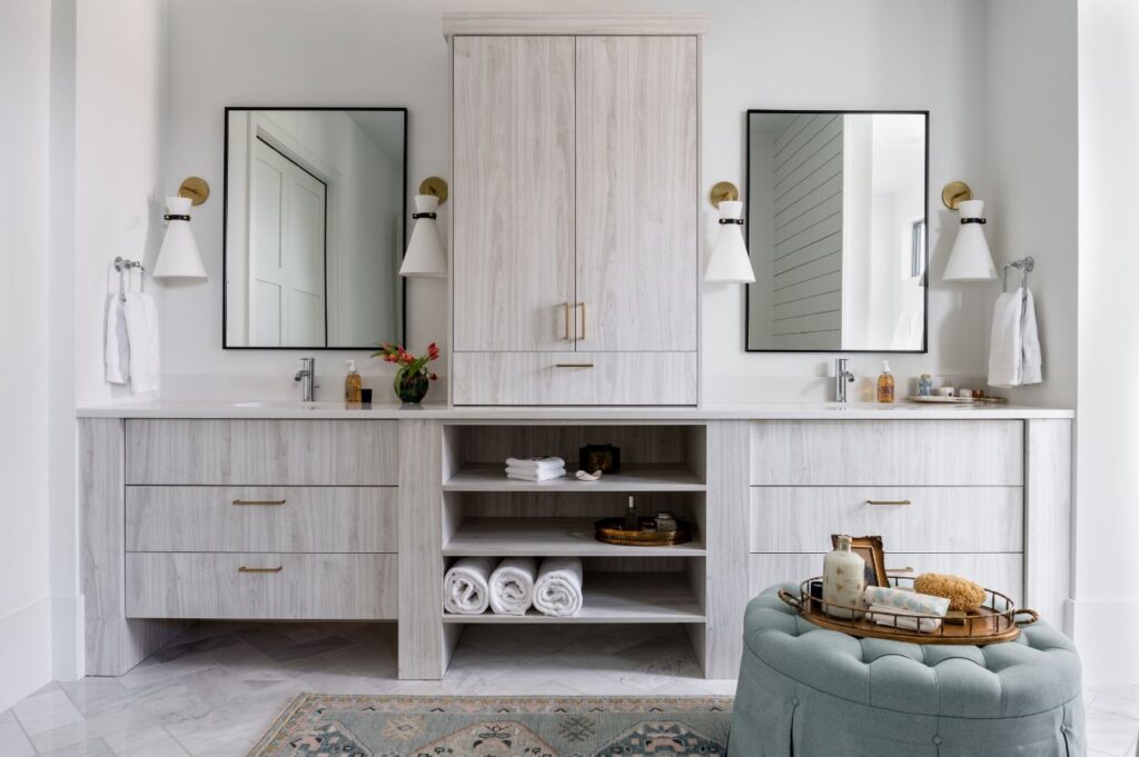 Modern bathroom by an interior designer Boerne TX, featuring dual sinks, two mirrors, white cabinetry, gold accents, open shelves with towels, and wall-mounted lights. A round ottoman and spa tray rest on a patterned rug for a stylish touch.
