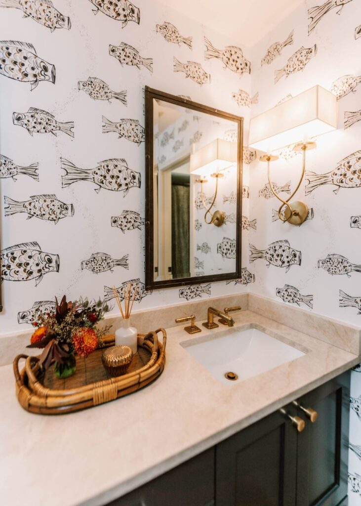 Bathroom with a marble countertop, gold fixtures, a rectangular mirror, and fish wallpaper. Styled by an interior designer Boerne TX, it features wall-mounted lights and a wicker tray with flowers and a candle on the counter.