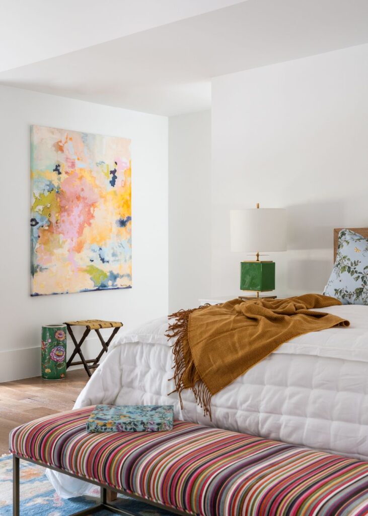 A bright bedroom styled by an interior decorator in Boerne features a white quilted bed, mustard throw, green lamp, floral pillow, and striped bench. An abstract painting and small stool add colorful accents to this cozy, modern space.
