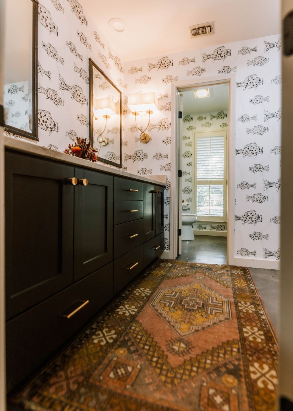 A bathroom by an interior designer Boerne TX features black cabinets, gold hardware, a patterned rug, and fish-patterned wallpaper. A double light fixture and mirror sit above the counter, with a doorway to a bright room with large windows and plantation shutters.