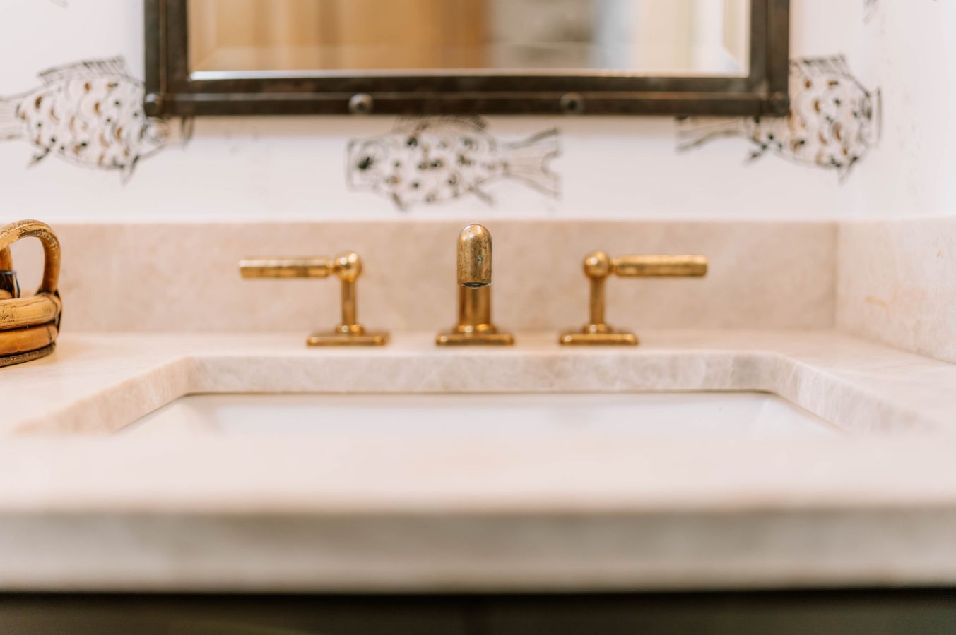 Close-up of a bathroom sink with marble countertop and gold faucet. A mirror and fish-illustrated wallpaper complete the look, showcasing the elegant touch of an interior decorator Boerne homeowners trust.