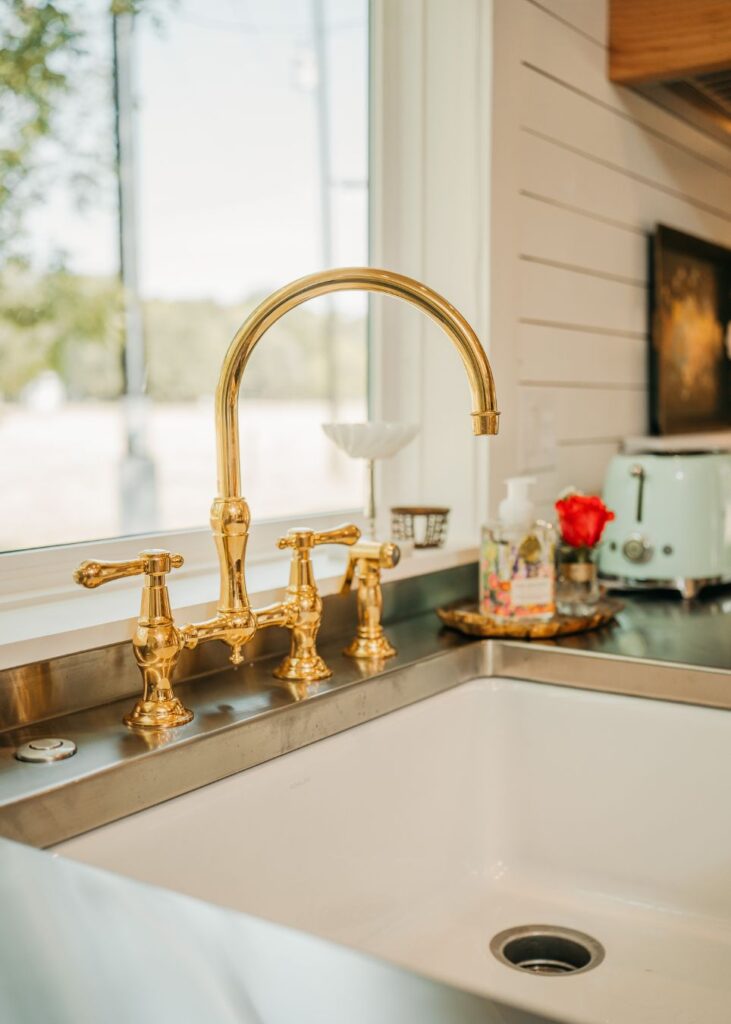A close-up of a gold kitchen faucet with three handles above a white sink, styled by an interior designer Boerne TX. Nearby are a soap dispenser, red flowers, and a mint-green toaster on the counter against a window.
