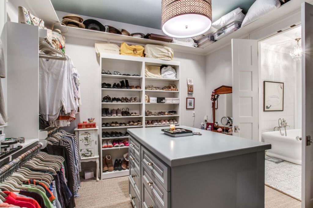 A spacious walk-in closet designed by an interior designer Boerne TX features shelves of shoes, hanging clothes, a dresser island, and bags on high shelves. An open door reveals a bathroom with a bathtub and framed art.
