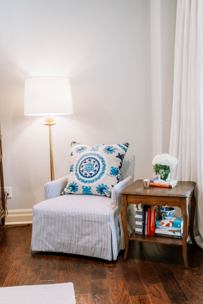 A cozy reading nook designed by an interior decorator Boerne features a striped armchair, blue patterned pillow, wooden side table with books, white flowers, and a lit lamp on a warm wooden floor.