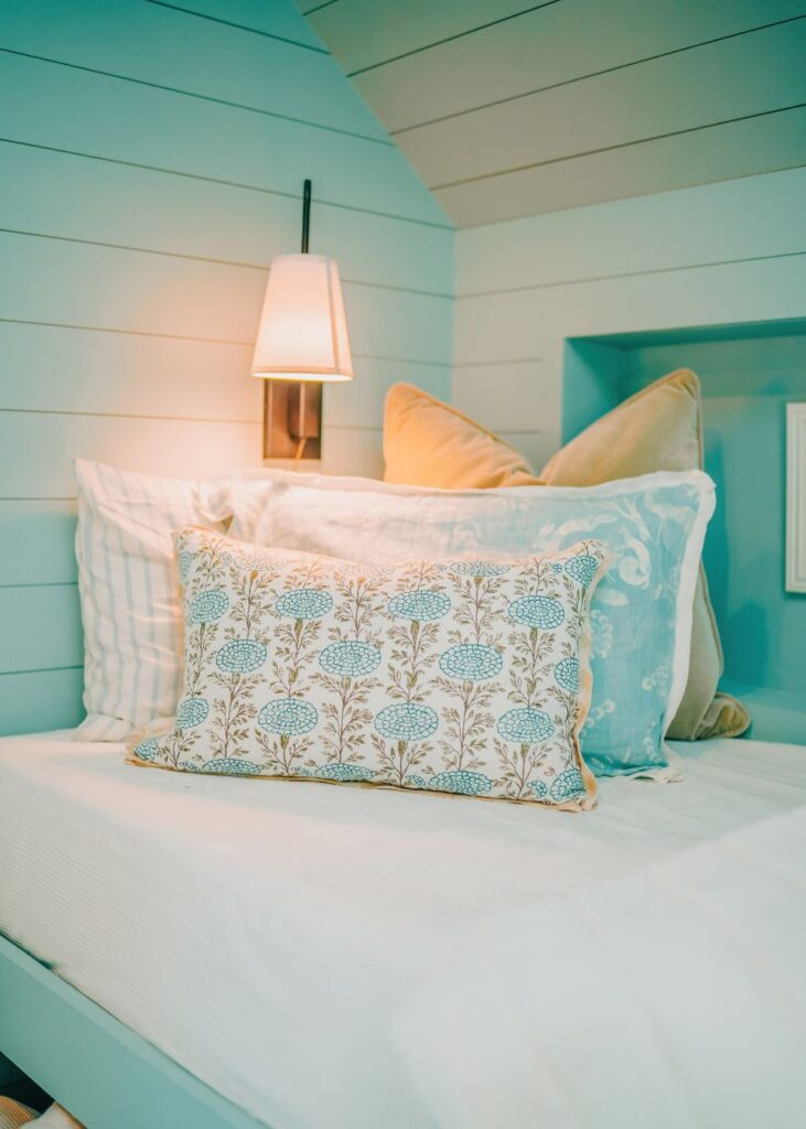 A cozy bed with white bedding, pastel blue and beige accent pillows, and a blue floral decorative pillow. Light wood-paneled walls and a wall-mounted lamp create an inviting space inspired by interior design Boerne TX style.