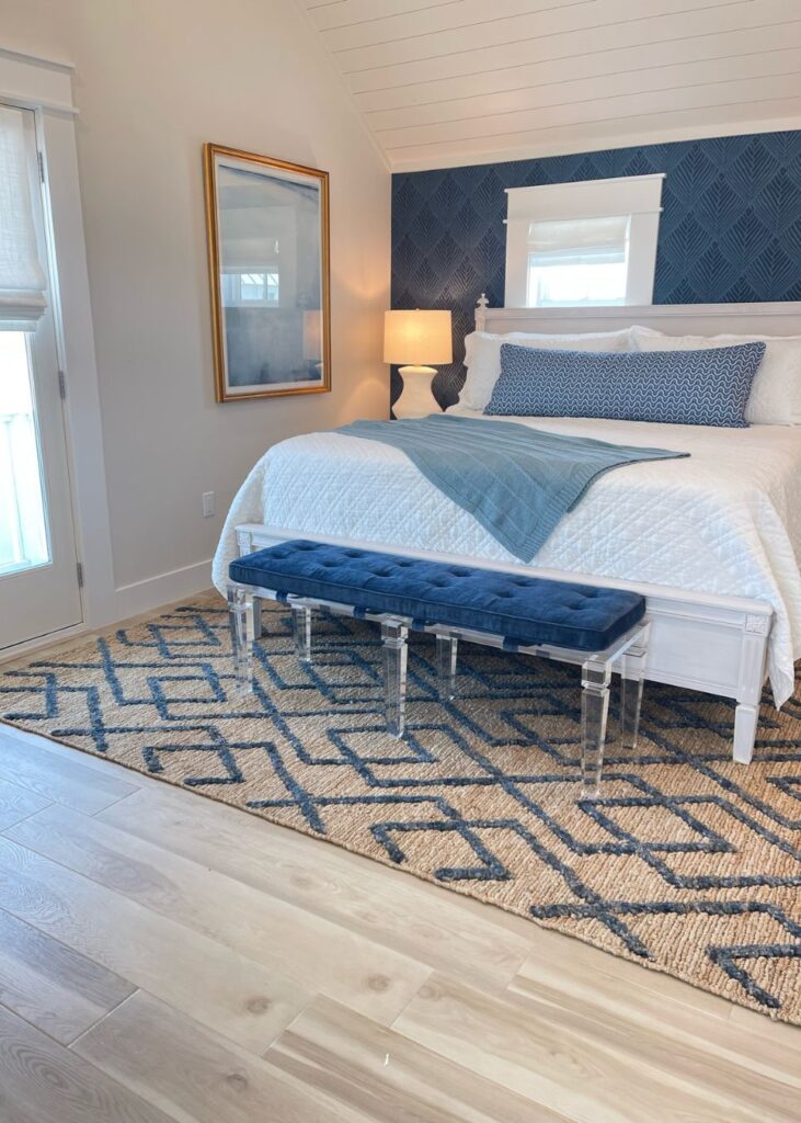 A modern bedroom with a white bed and blue accents, styled by an interior design company Boerne TX, features a navy suede bench, blue throw, geometric-patterned rug, lit table lamp, framed wall art, and a textured blue accent wall for warmth and style.