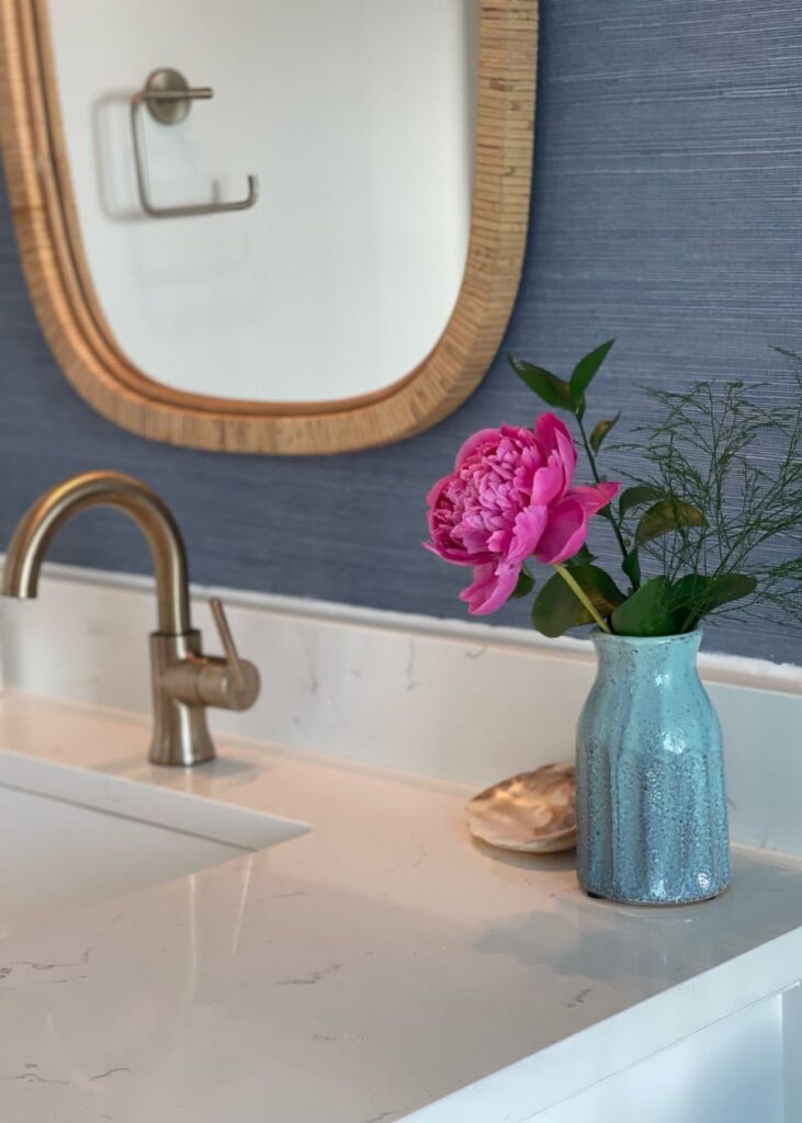 A bathroom sink with a brass faucet, a blue vase holding a pink peony and greenery, a seashell, and a mirror with a wooden frame on a blue wall styled by an interior designer Boerne TX.