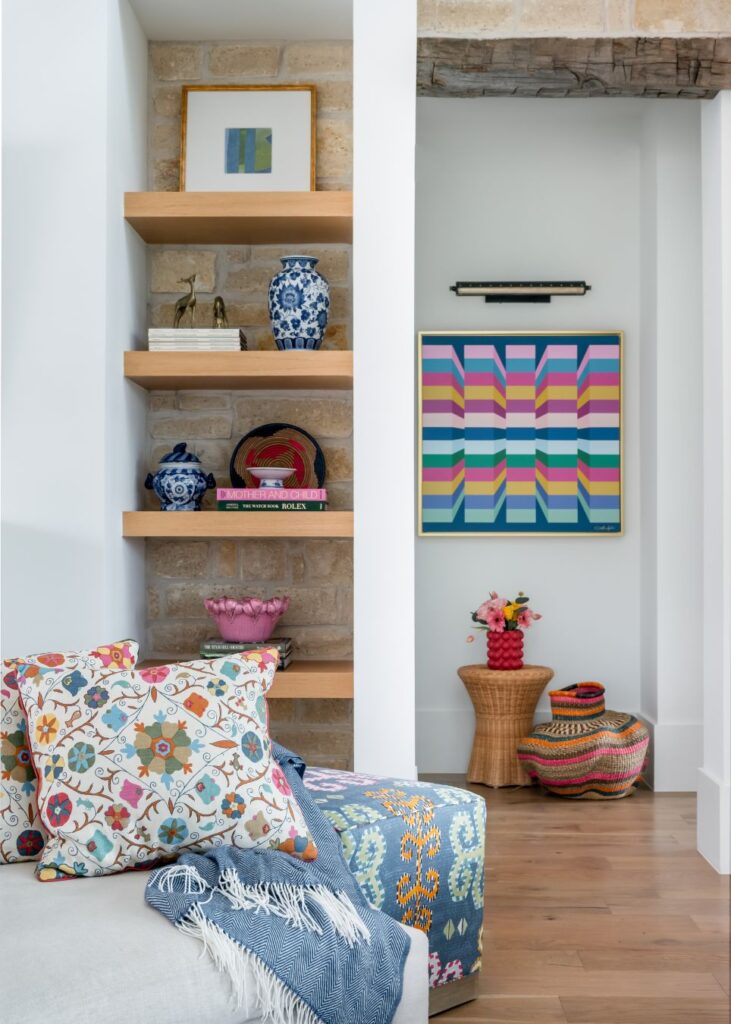 A cozy living space styled by an interior design company in Boerne, TX, featuring patterned pillows, wood shelves with blue vases, and vibrant geometric wall art above woven baskets and stools on a wooden floor.