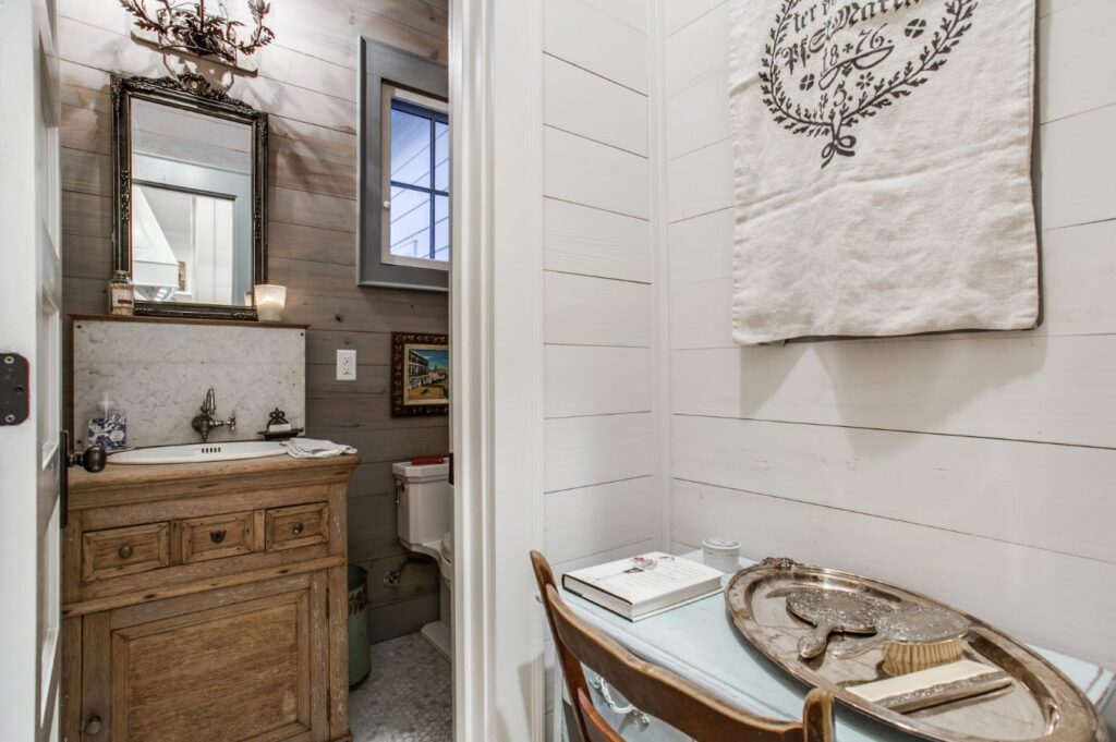A rustic bathroom with wood-paneled walls, a wooden vanity with a sink and mirror, a toilet, and a small corner desk; styled by an interior decorator Boerne, it features books, a decorative tray, and a wall hanging above the desk.