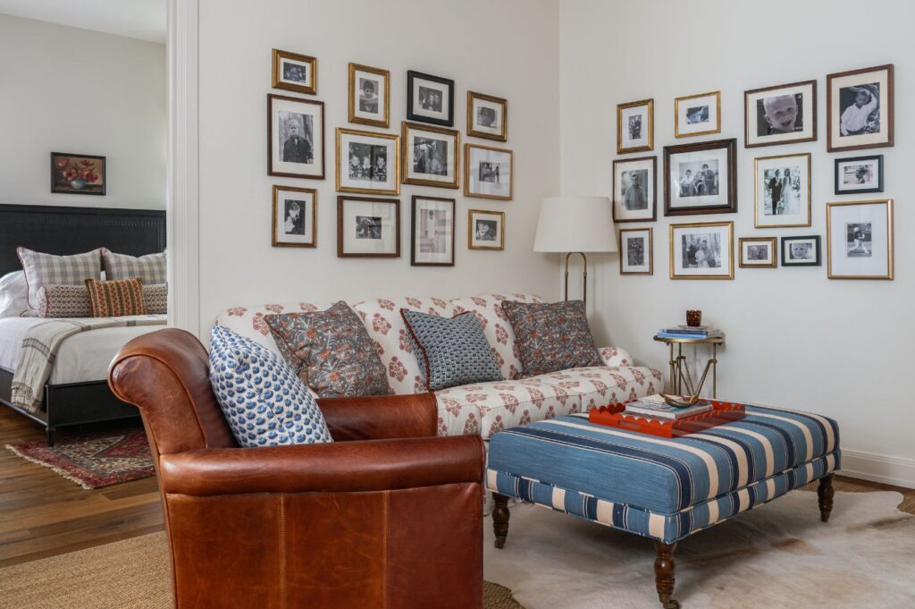 A cozy living room designed by a historic home interior designer features a brown leather chair, a floral sofa with patterned pillows, a blue-striped ottoman, and walls adorned with black-and-white photo frames; a bedroom is visible in the background.